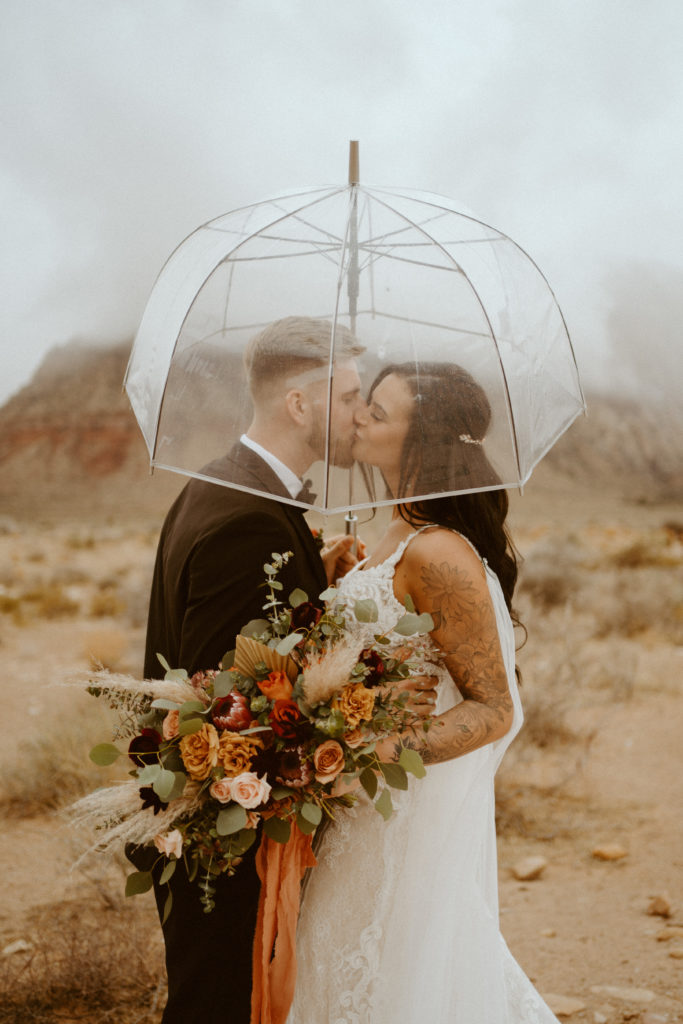 Bride with fall inspired bouquet with Groom kissing under transparent umbrella for Red Rock Bridals & Simple Affair Micro-Wedding 