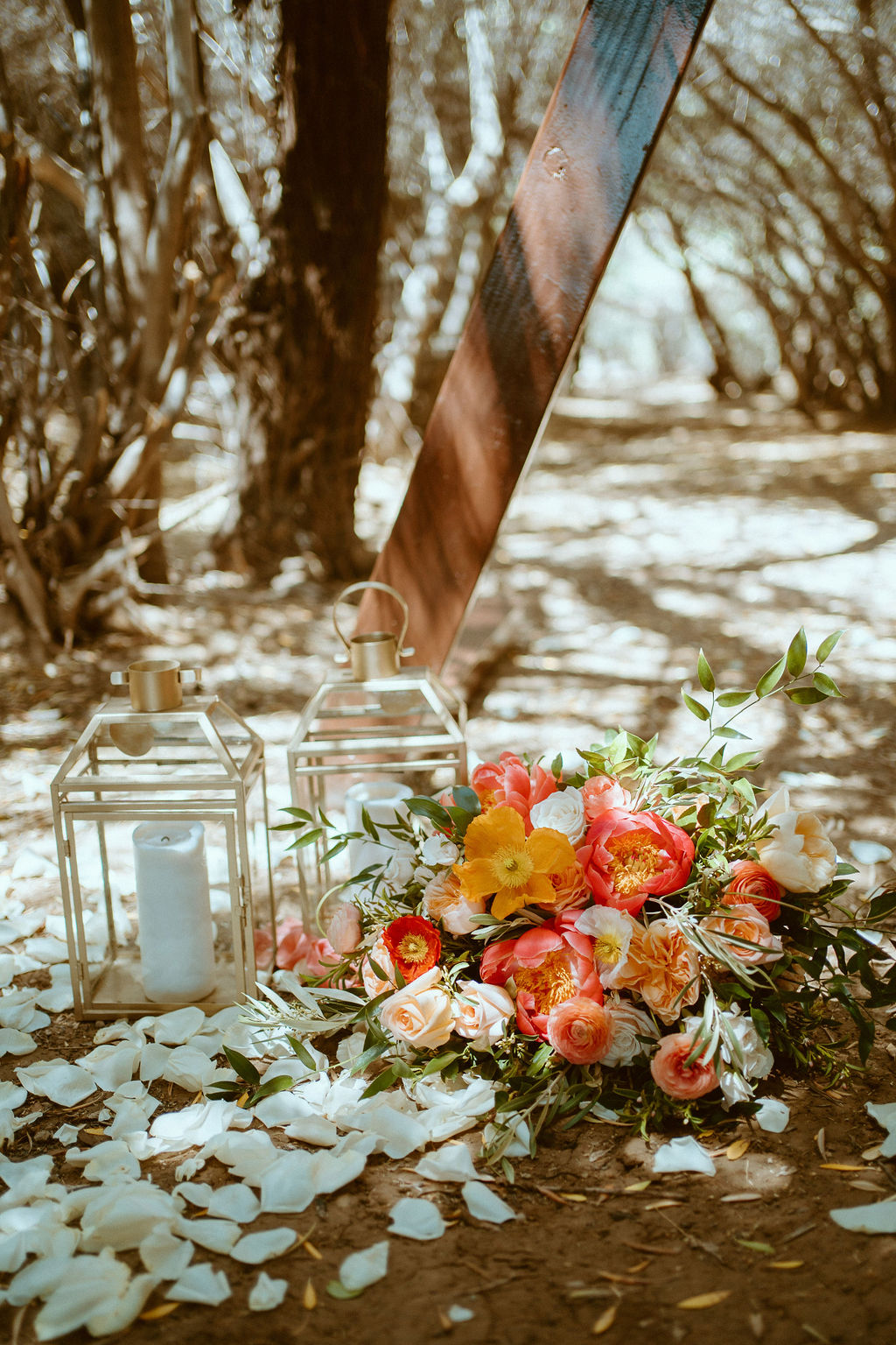 two lanterns on the ground with flower pedals next to arch with brides bouquet 