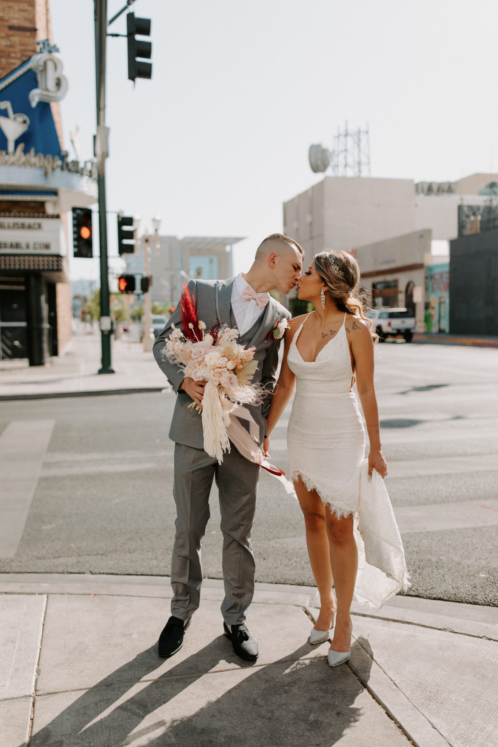 Groom in grey suit and pink bowtie holding bride's bouquet and kissing bride in scoop neck lace bridal dress while walking in Las Vegas 