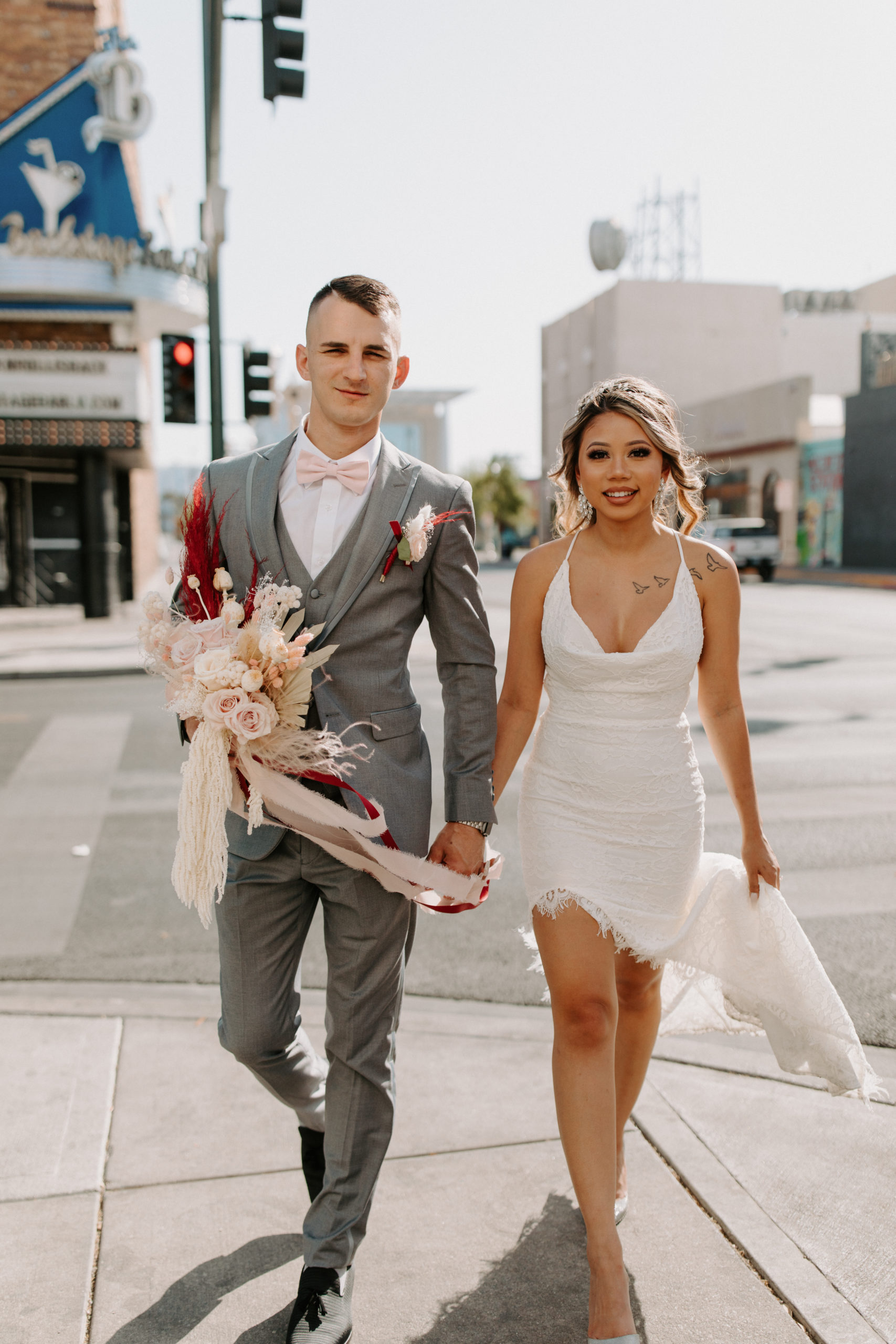 Groom in grey suit and pink bowtie holding bride's bouquet with bride in scoop neck lace bridal dress while walking in Las Vegas 
