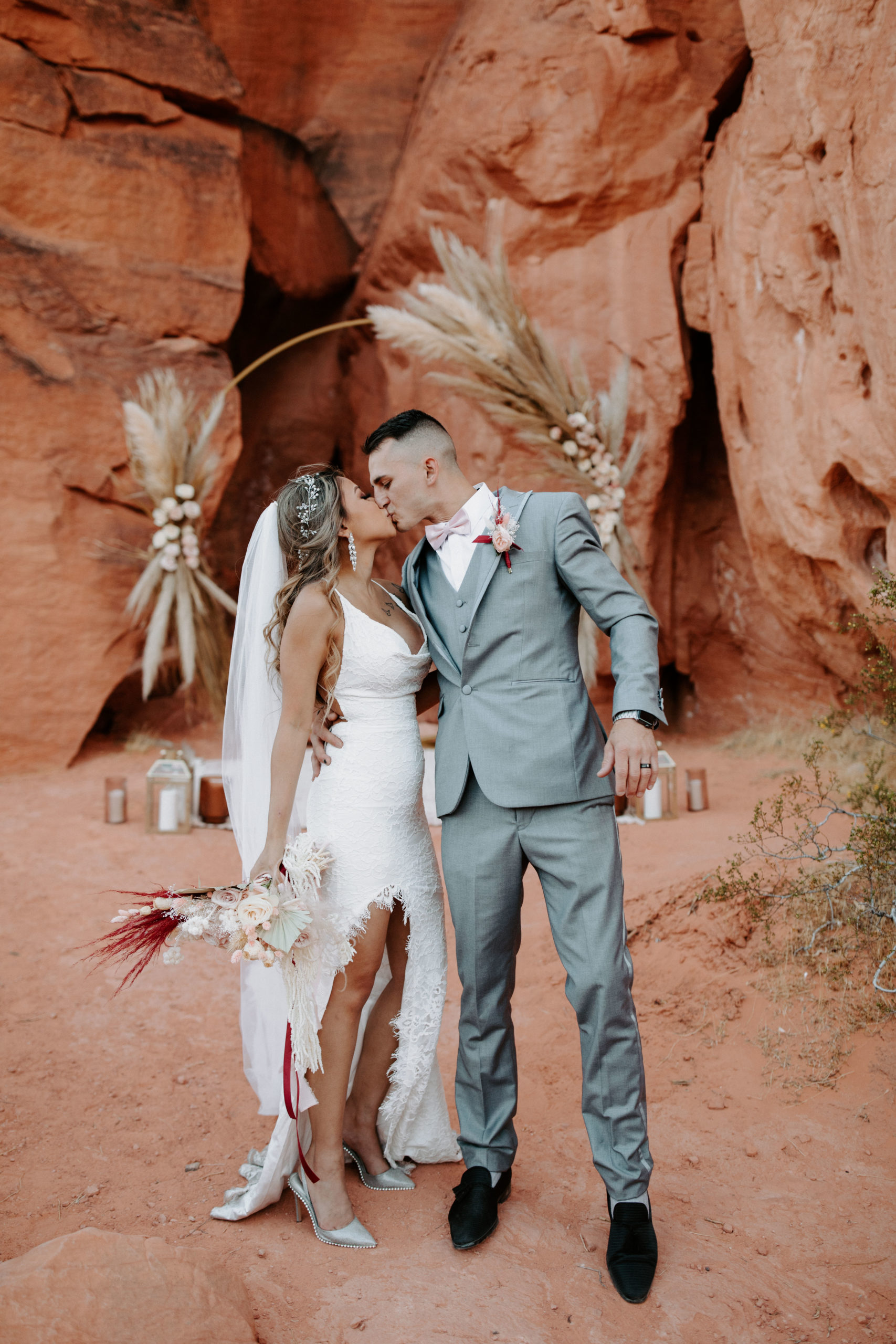 Newlyweds kissing after Boho Pampas Desert Elopement. Groom wearing gray suit with pink bowtie and bride in lace bridal gown with leg slit for monochromatic light pink theme with red accent  