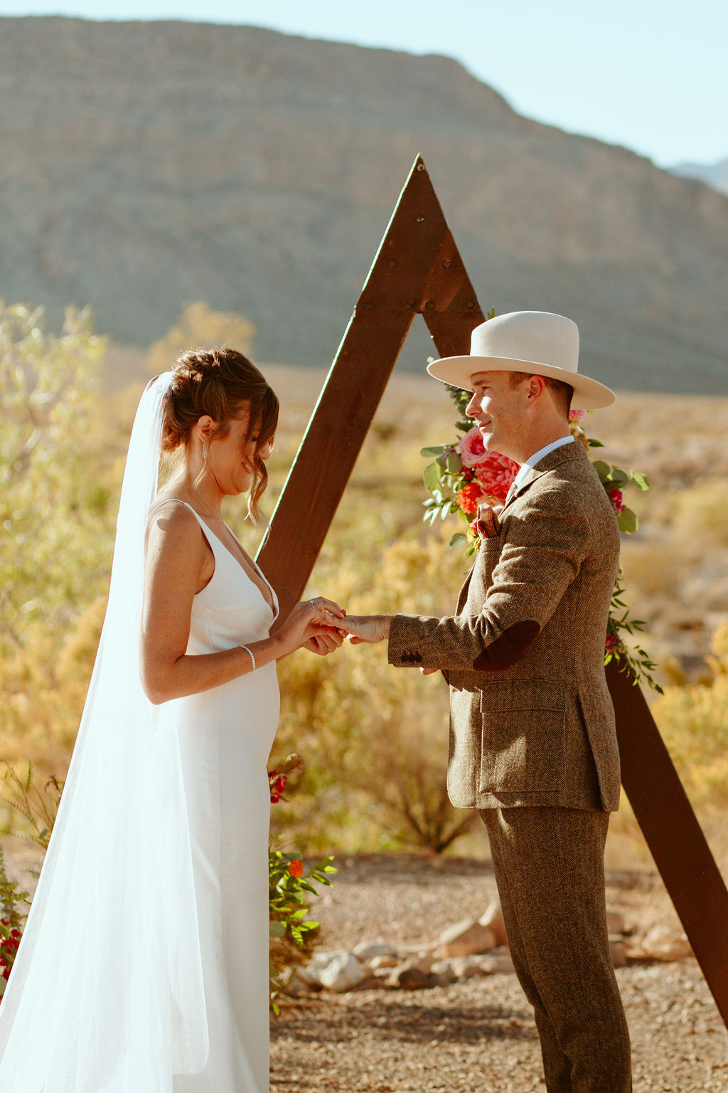 Groom in large brim fedora hat and bride in white gown with long veil  putting rings on each others fingers 
