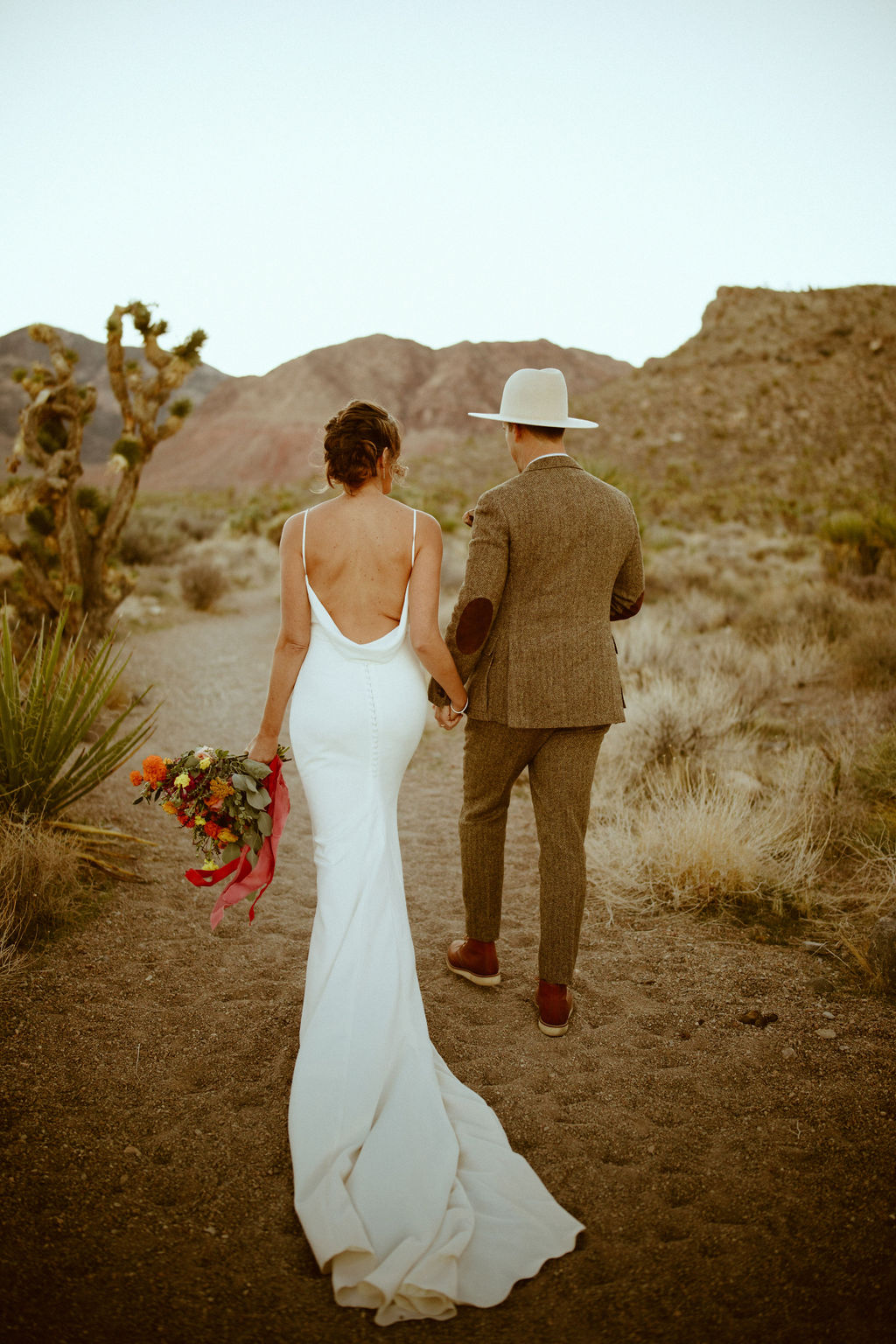 Bride in white backless gown with Groom in brown tweed suit and cream colored hat walking through desert together with cactus and mountains 