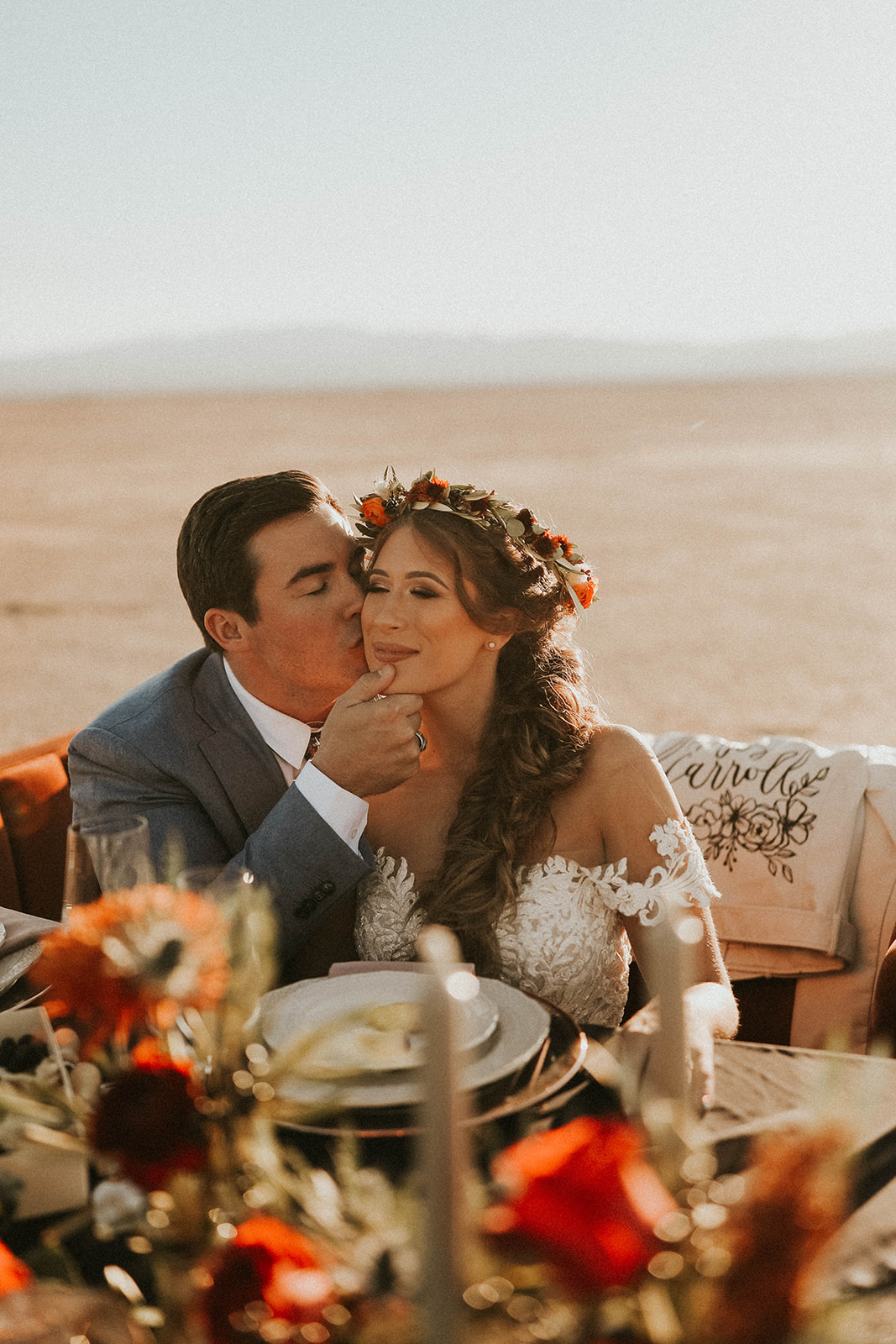 Groom Kissing Bride's Cheek while Sitting at Dry Lake Bed Fall Inspired Elopement Sweetheart Table with Velvet Couch & Fall Inspired Floral 