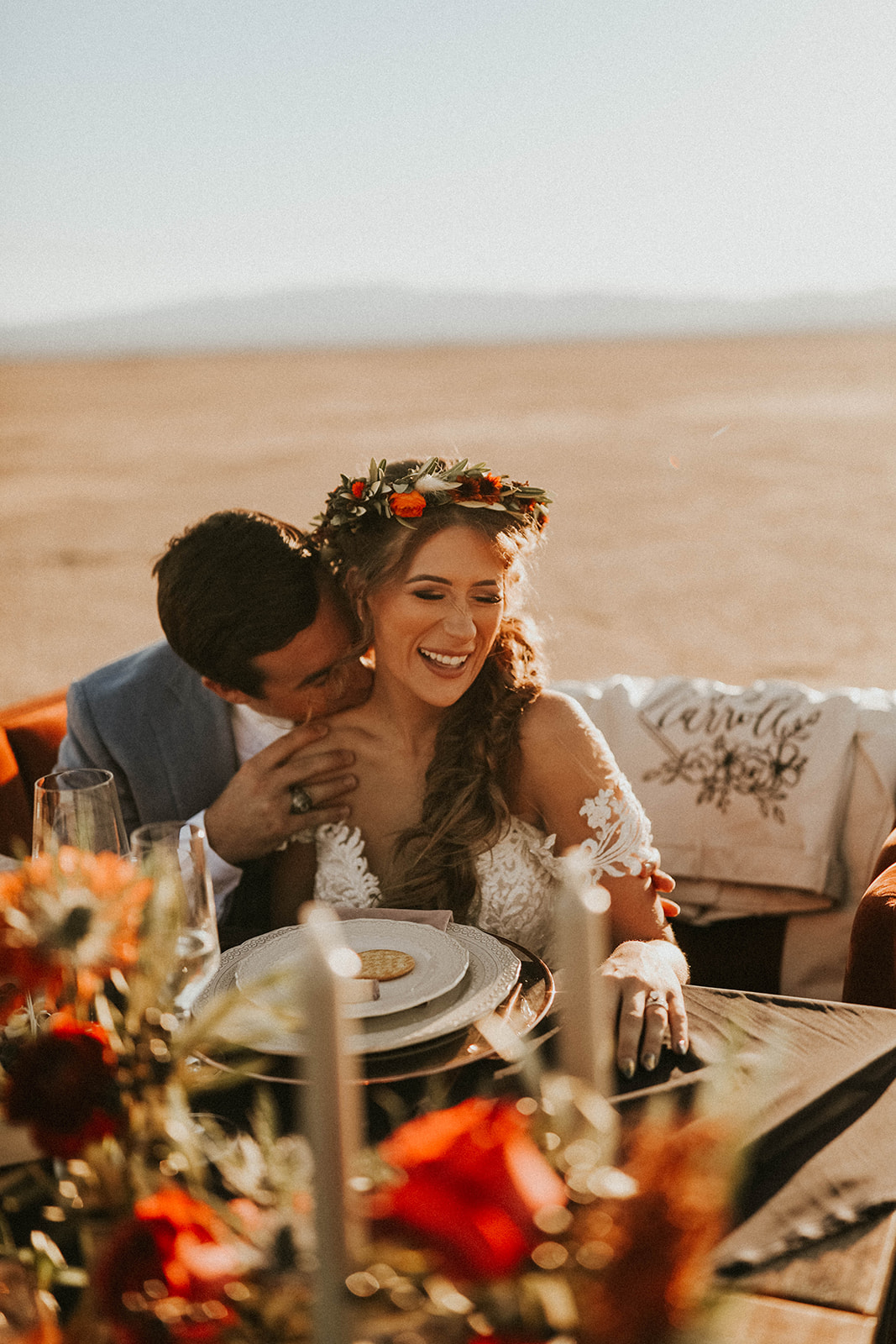 Groom Kissing Bride's Shoulder while Sitting at Dry Lake Bed Fall Inspired Elopement Sweetheart Table with Velvet Couch & Fall Inspired Floral 