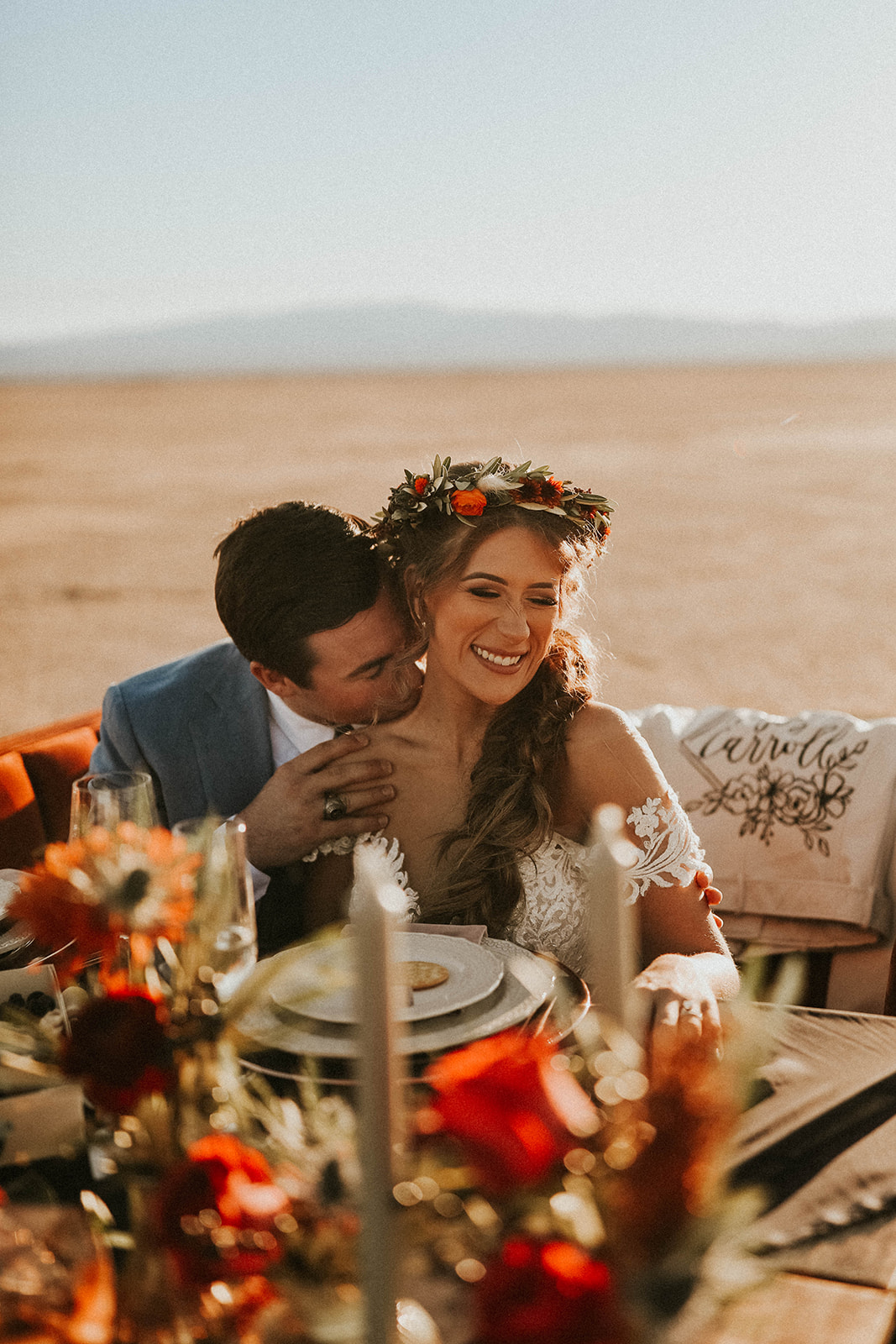 Groom Kissing Bride's Shoulder while Bride Smiles Sitting at Dry Lake Bed Fall Inspired Elopement Sweetheart Table with Velvet Couch & Fall Inspired Floral 