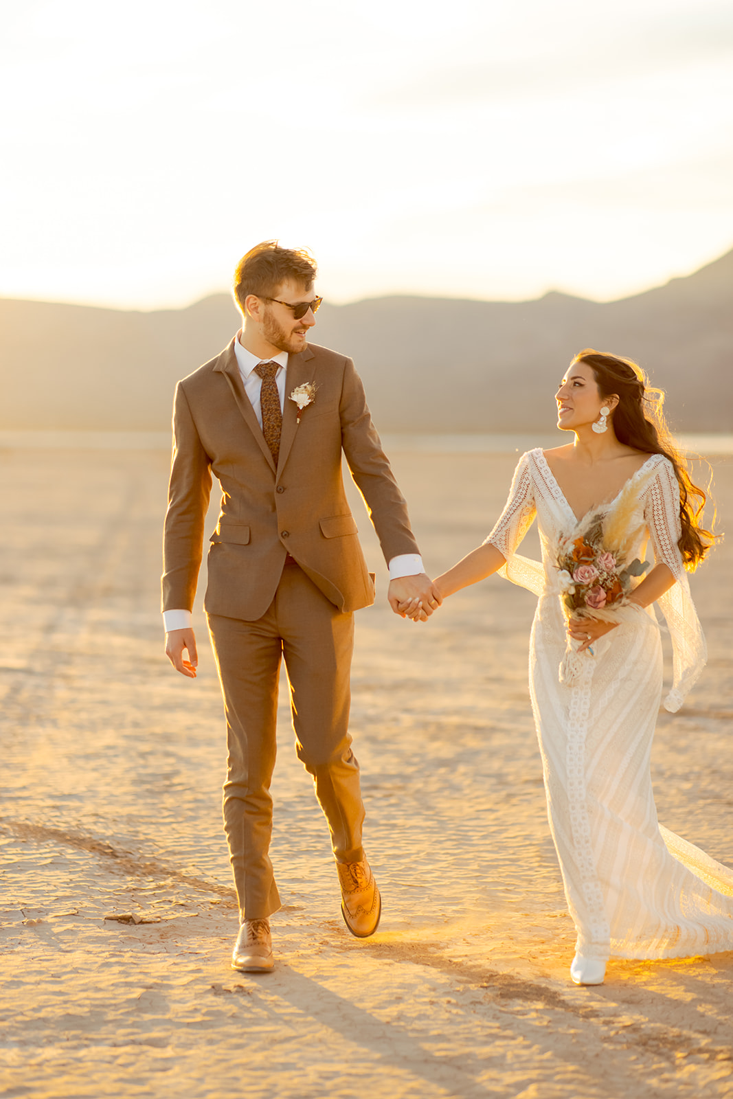 Bride in long sleeved boho dress walking with groom in vintage brown suit during sunset for Dry Lake Bed Micro-Wedding