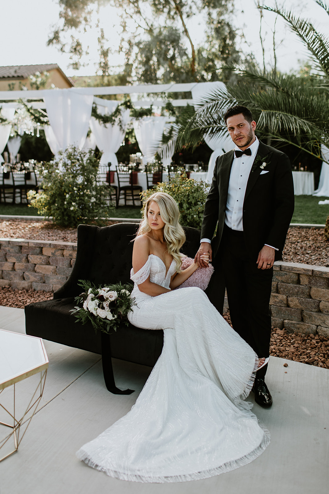 Bride on Black Velvet Couch holding hands with Groom with Moody Modern Glam Outdoor Reception Set up behind them at Lotus House 