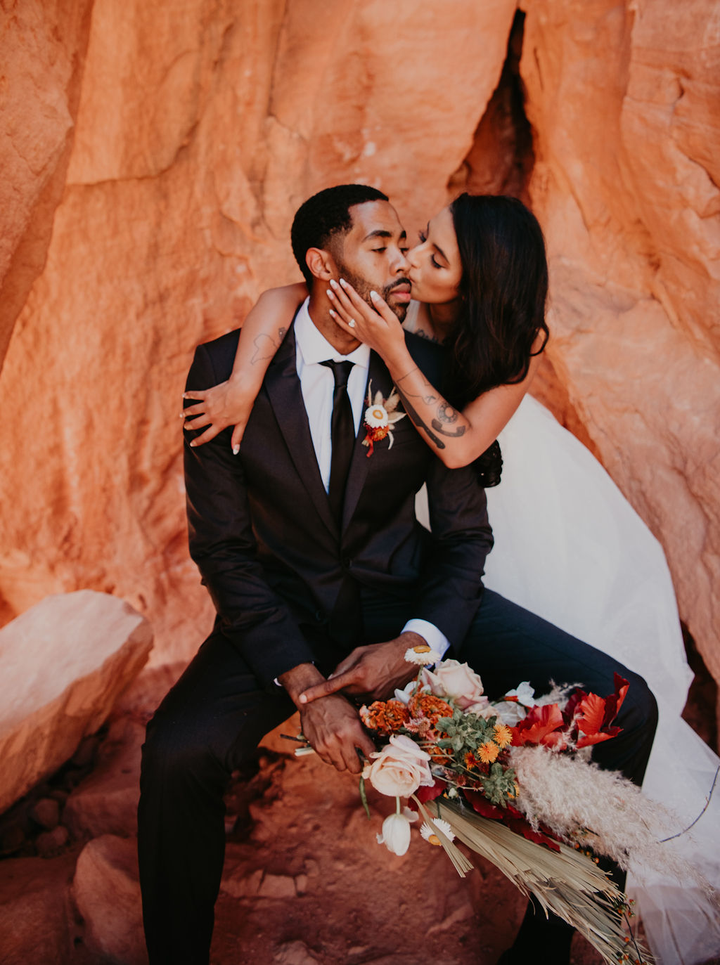 Bride Kissing Grooms Cheek while Groom is Sitting on Red Rock at Valley of Fire 