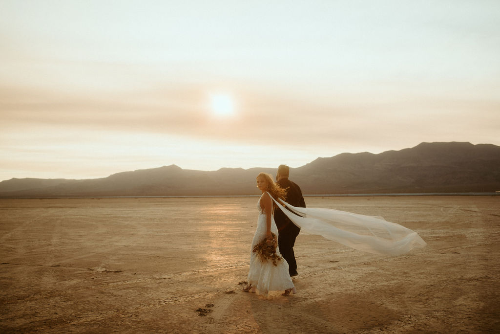 Bride with Bouquet and Bridal Cape Walking in Desert with Groom 