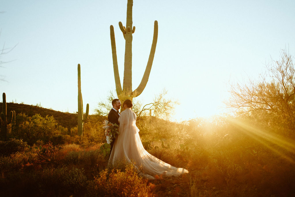 Saguaro National Park Micro-Wedding. The groom holding the bride closely and she hangs down her bouquet. Standing in front of a cactus with the beautiful sunset beaming behind them