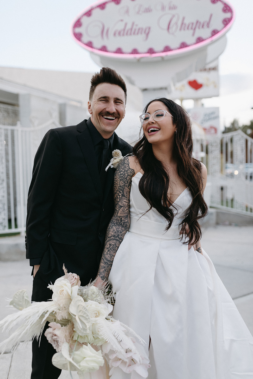 Groom in All Black Wedding Suit and Bride with Clear Statement Glasses and White Dress Outside Vegas Chapel 
