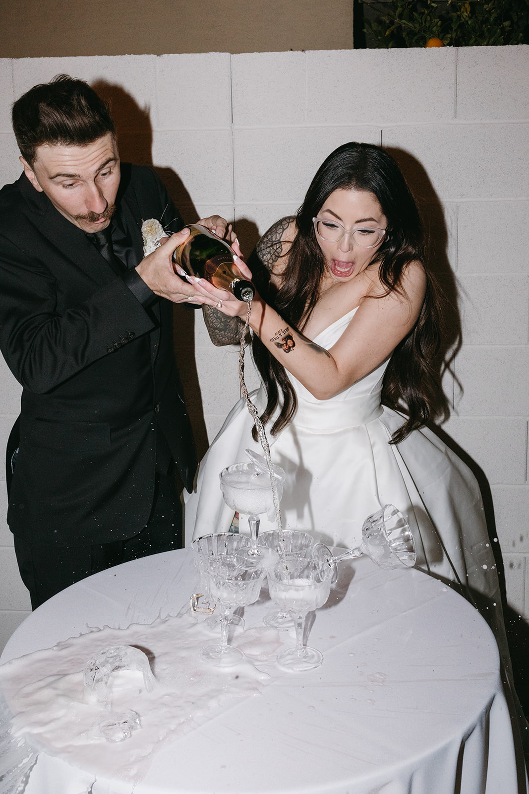 Couple spilling and pouring Champagne in Champagne glass tower during their fun backyard reception dinner 