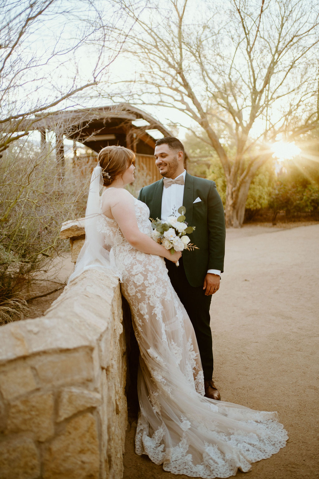 Bride leaning on rock wall with groom during sunset 