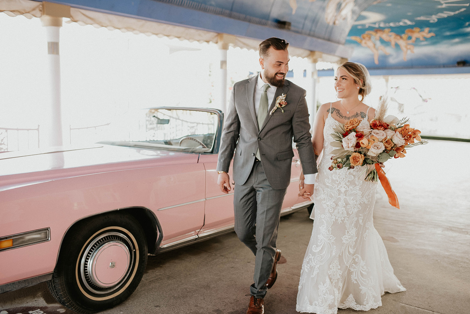 First Stop! A Little White Chapel Bride and groom holding hands in front of a pink Cadillac. Lace thin strap bridal gown with boho style bouquet. Groom in a dark grey suit with boho boutonniere pined to his lapel. 