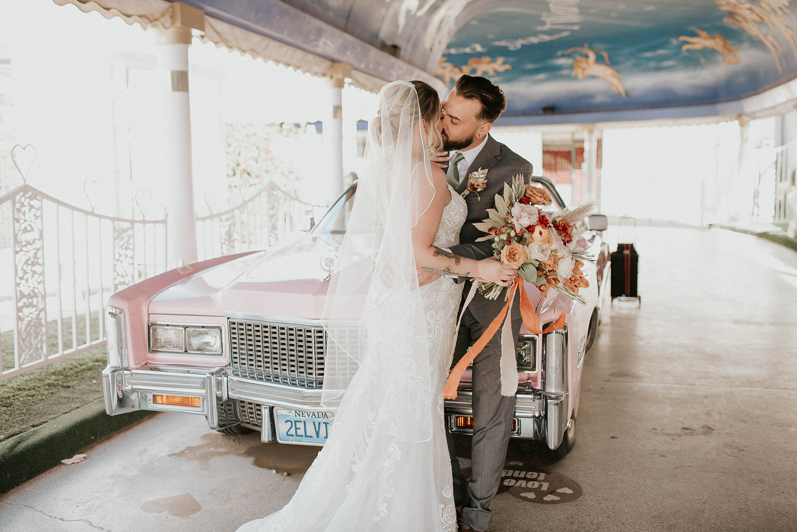 First Stop! A Little White Chapel sharing a kiss at the front of bright pink Cadillac. Veil cascading down the brides soft curled updo. Orange ribbon flowing in wind with boho styled bouquet. 