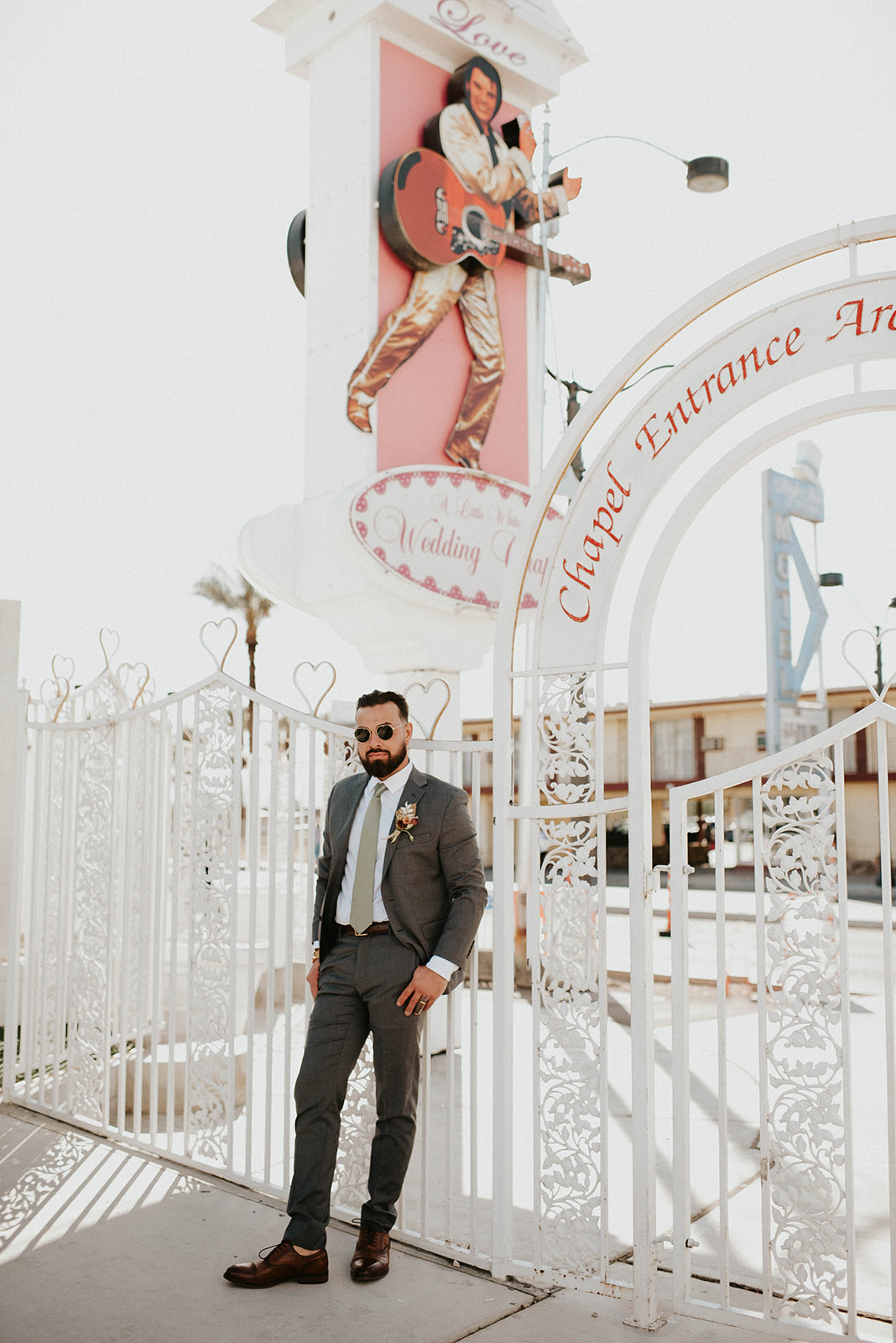 Groom wearing sunglasses leaning against the white gates at the Little White Chapel. Elvis sign in the background. 