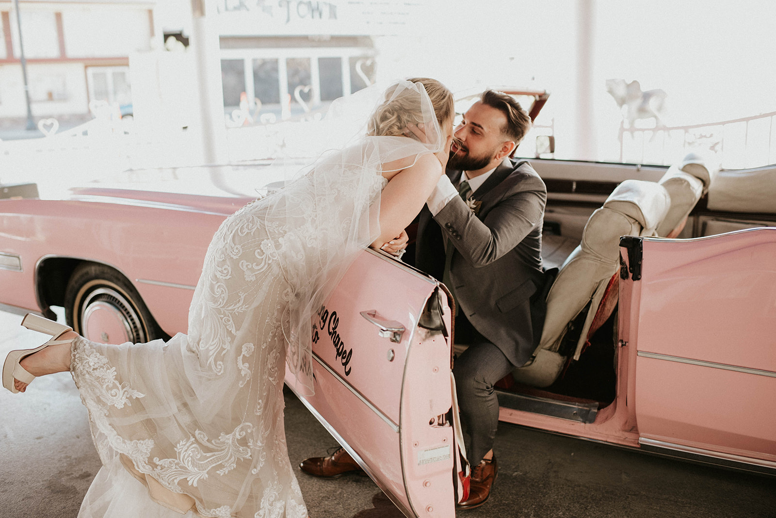 First Stop! A Little White Chapel Bride leans over the pink Cadillac kissing the groom. Kicking her foot out with her cute white chunky heals. 