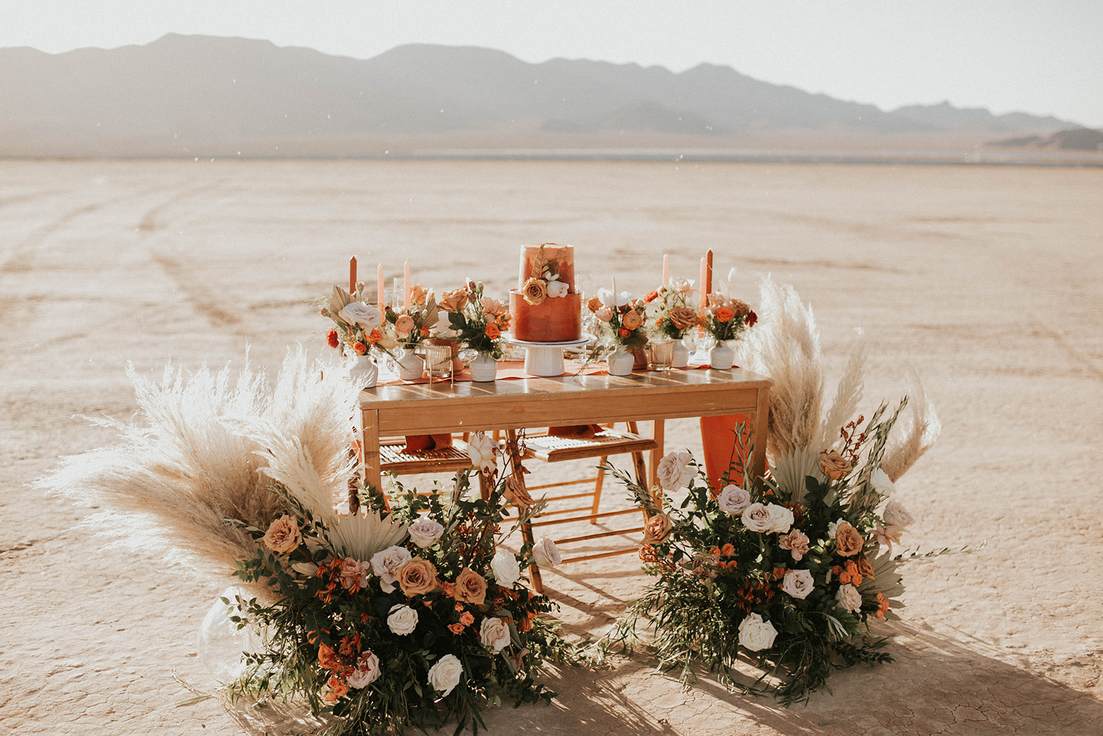 First Stop! A Little White Chapel sweetheart table decorated with bright orange boho styled florals and two tiered cake. Two ground floral arches decorate each side of the table. A deep burnt orange runner lays across the wooden table. 