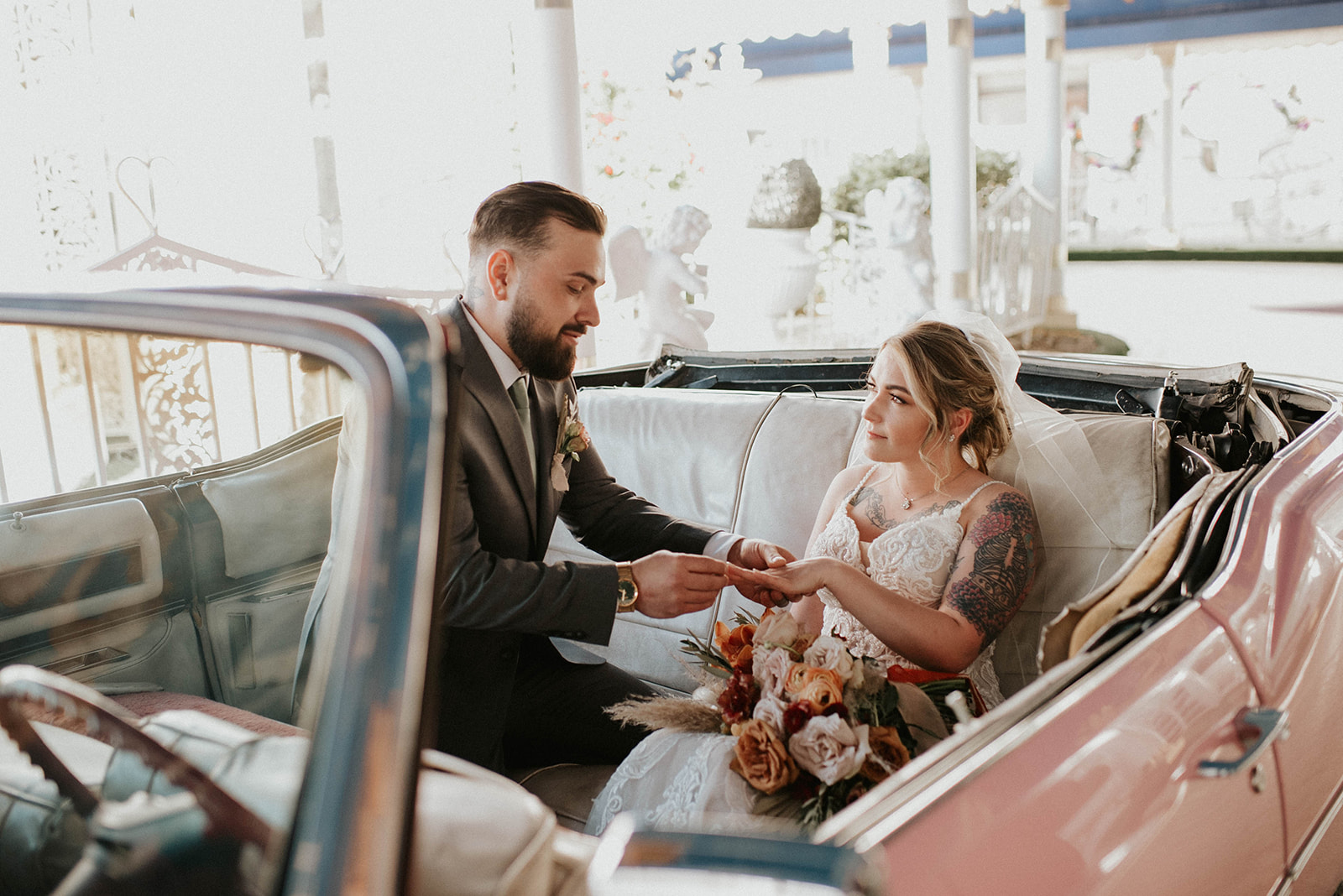 First Stop! A Little White Chapel the groom places the ring on the brides hand sitting inside a pink Cadillac. Boho bouquet resting in lap. A soft low updo with thin veil in her hair. 