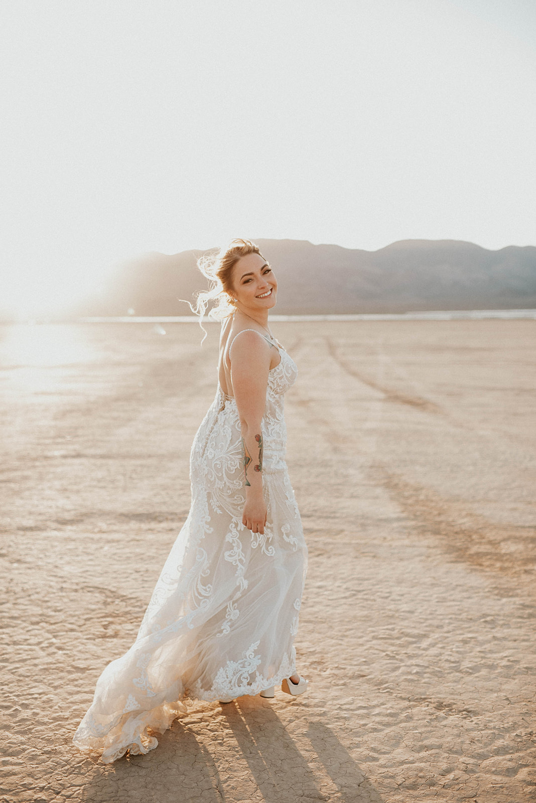 First Stop! A Little White Chapel bride walking in the desert with a beautiful smile the sunset right behind her. The bride wears a lace fitted bridal gown with buttons down the back and thin straps. The gown has a small train behind it. 
