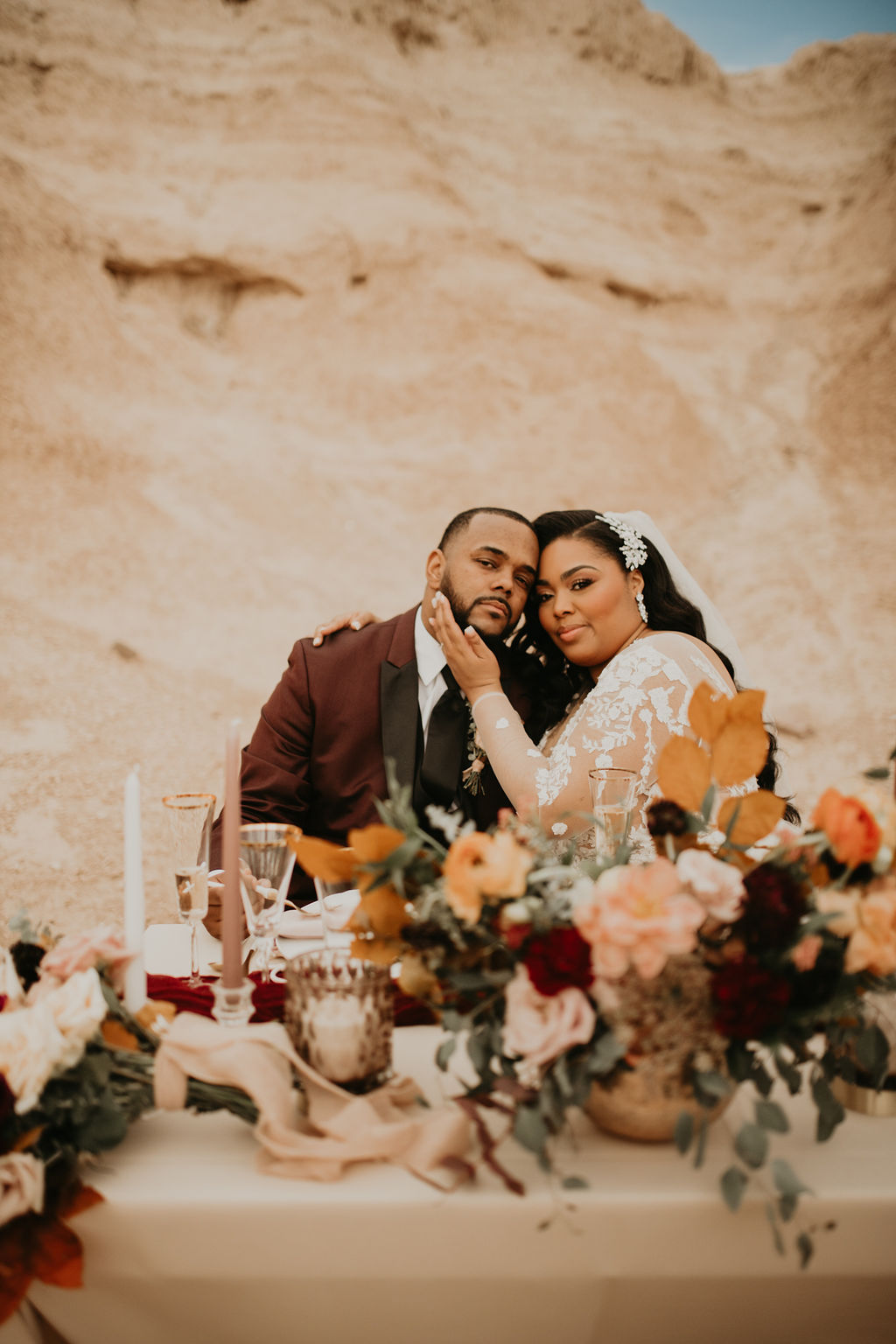Newlyweds sitting behind their gold and burgundy sweetheart table. The bride and groom have their heads leaning against each other while the bride has her hand placed softly on the grooms face.