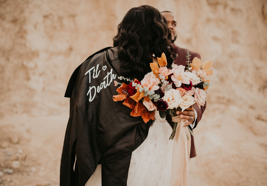 Fossil Beds Sunset Elopement the bride has her custom leather wedding jacket that says "till death" draped over her lacey wedding gown. 