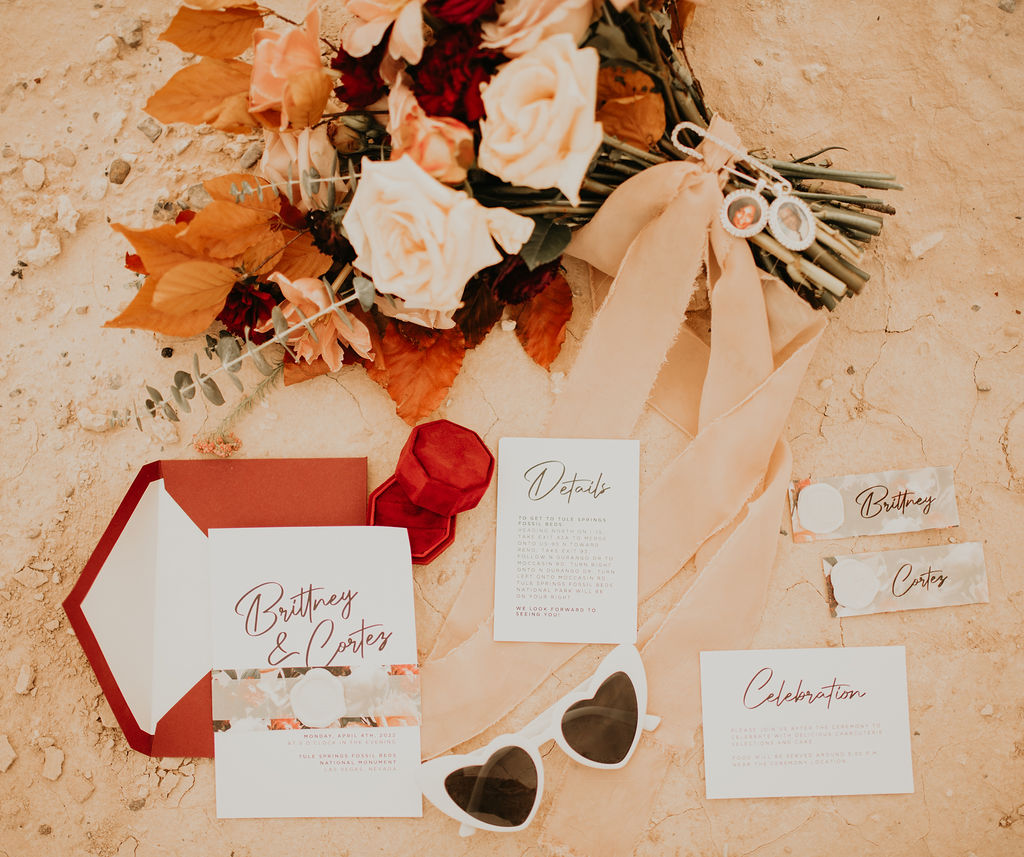 Invitation suite flat lay with the brides deep red red box. The couples wedding invite with a details and celebration card along with the bride and groom name tags for the sweetheart table. Next to the brides bridal bouquet with two pendants of loved ones, ribbon, and heart shaped retro sunglasses. 