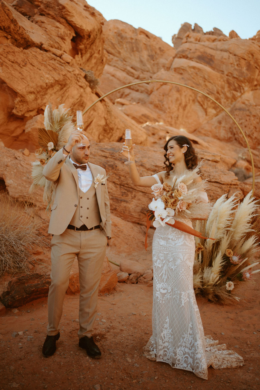 International Couples! Welcome to the desert! Bride holding boho bouquet with white orchids in front of circle arch with pampas grass florals. Couple doing champagne toast. 