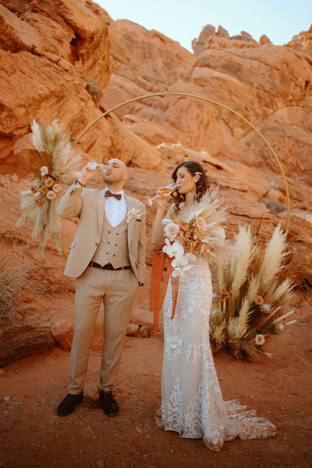 Groom in all tan suite and bowtie drinking champagne with bride in detailed lace dress for boho elopement. International Couples! Welcome to the desert!