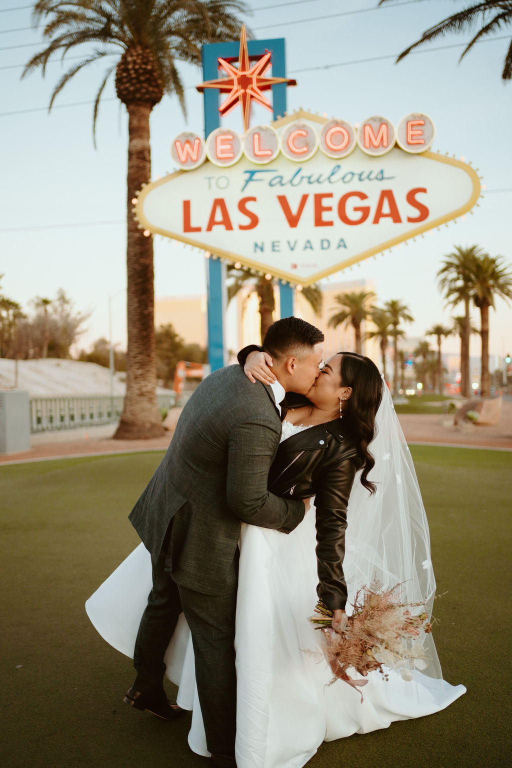 Bride with Dusty Rose bouquet and Custom Black Leather Bridal Jacket Kissing Groom in front Famous Las Vegas Sign during Las Vegas Highlights & Desert Elopement