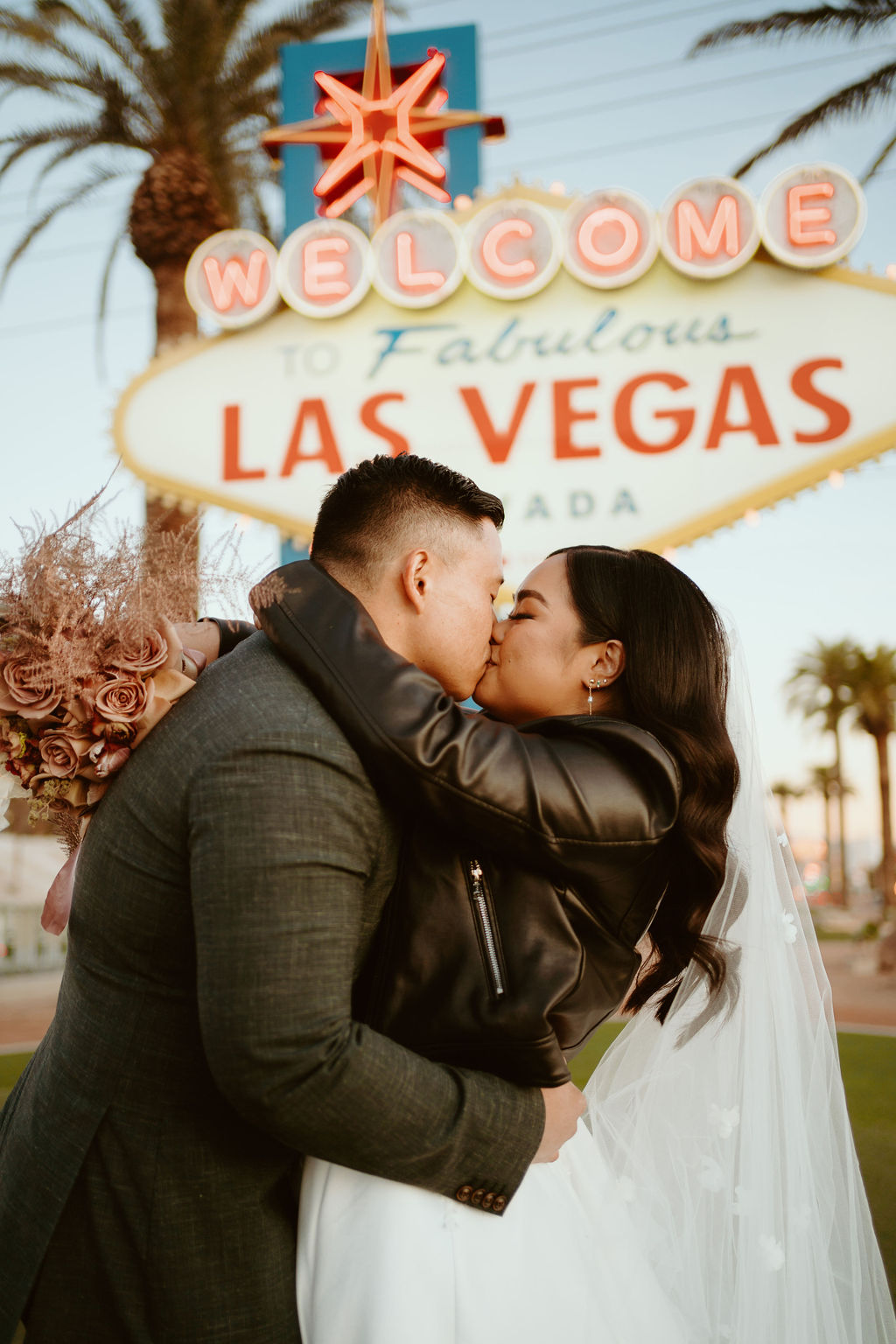 Bride with Dusty Rose bouquet and Custom Black Leather Bridal Jacket Kissing Groom in front of Welcome to Fabulous Las Vegas Nevada Sign