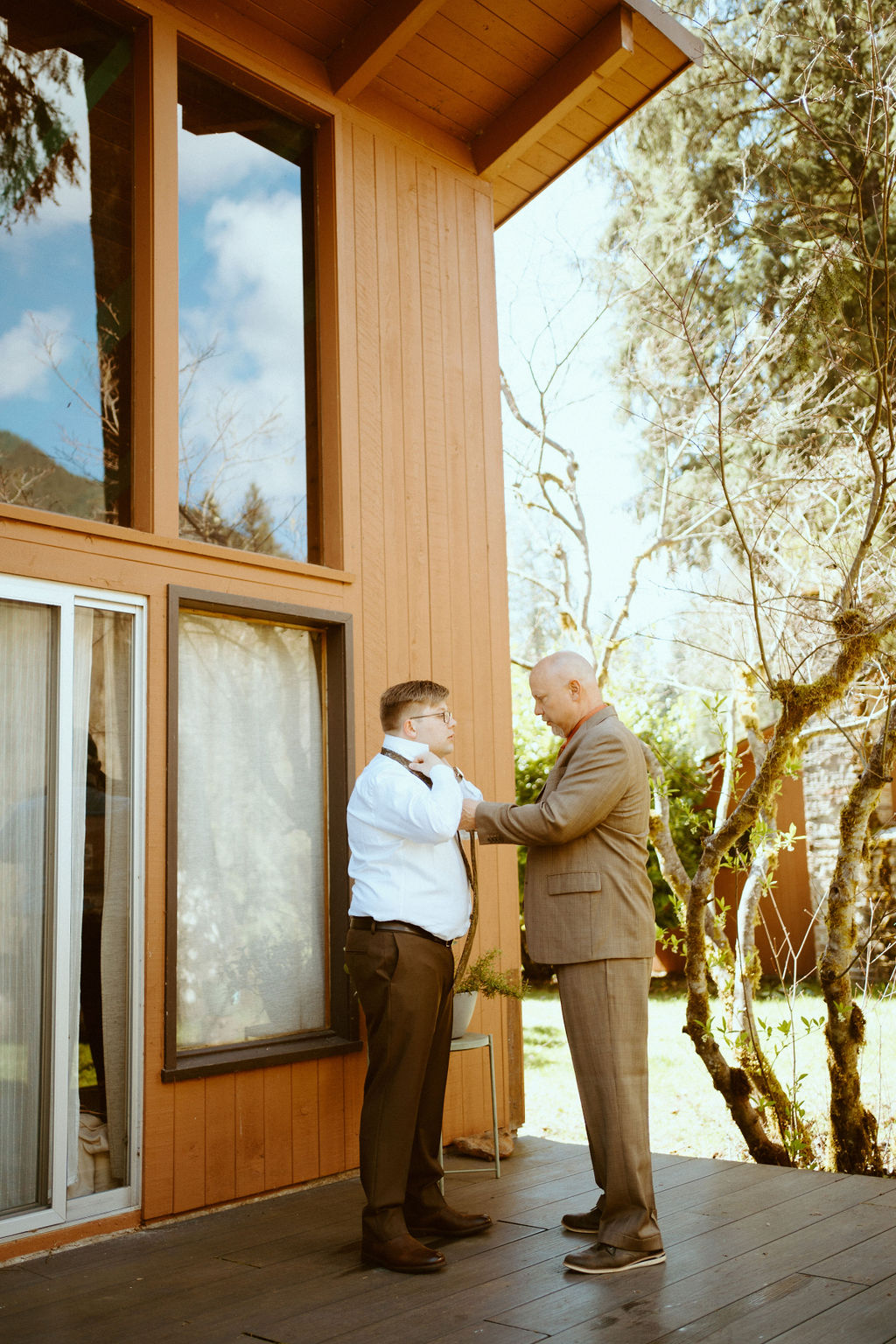 The grooms father is helping him get ready. Fixing his tie and sleeves of his button down shirt. Outside of the room on the balcony over looking the forest at Loloma Lodge.