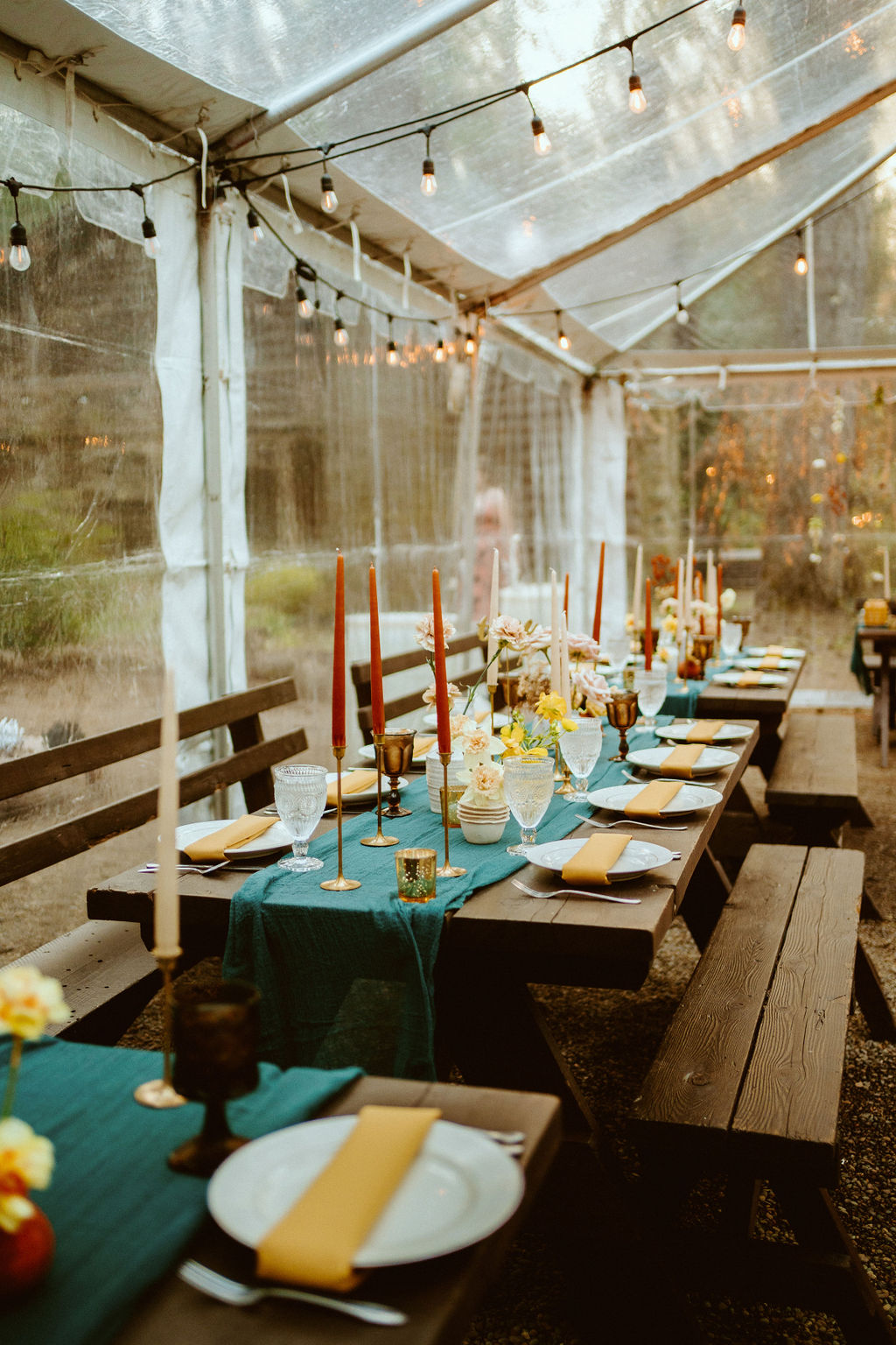 Guest tables that have a deep green table runner with small florals and tapper candles and wooden benches for seating