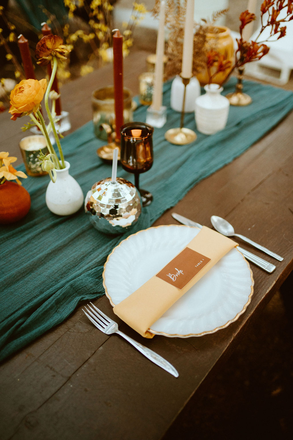 Place seating at the sweetheart table  with a white round plate and a gold trim and a creamy orange tri fold napkin on top of a wooden table with a turquoise table runner, small floral arrangements all over with rust and mustard taper candles, silver votives and amber gablets and disco ball cups. 