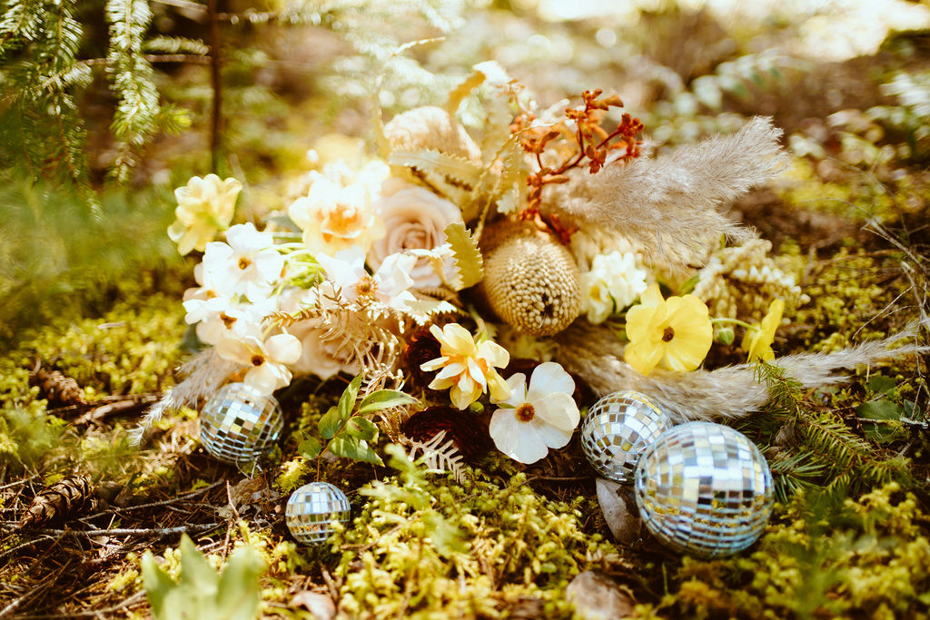 A floral piece with cosmos, roses, dried florals, pampas that are beautiful neutral colors with pops of yellow and amber mixed in with different size disco balls.