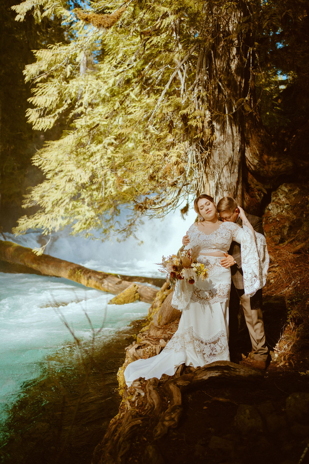 Couple posing on rocks while the groom kisses the bride in front of a waterfall 