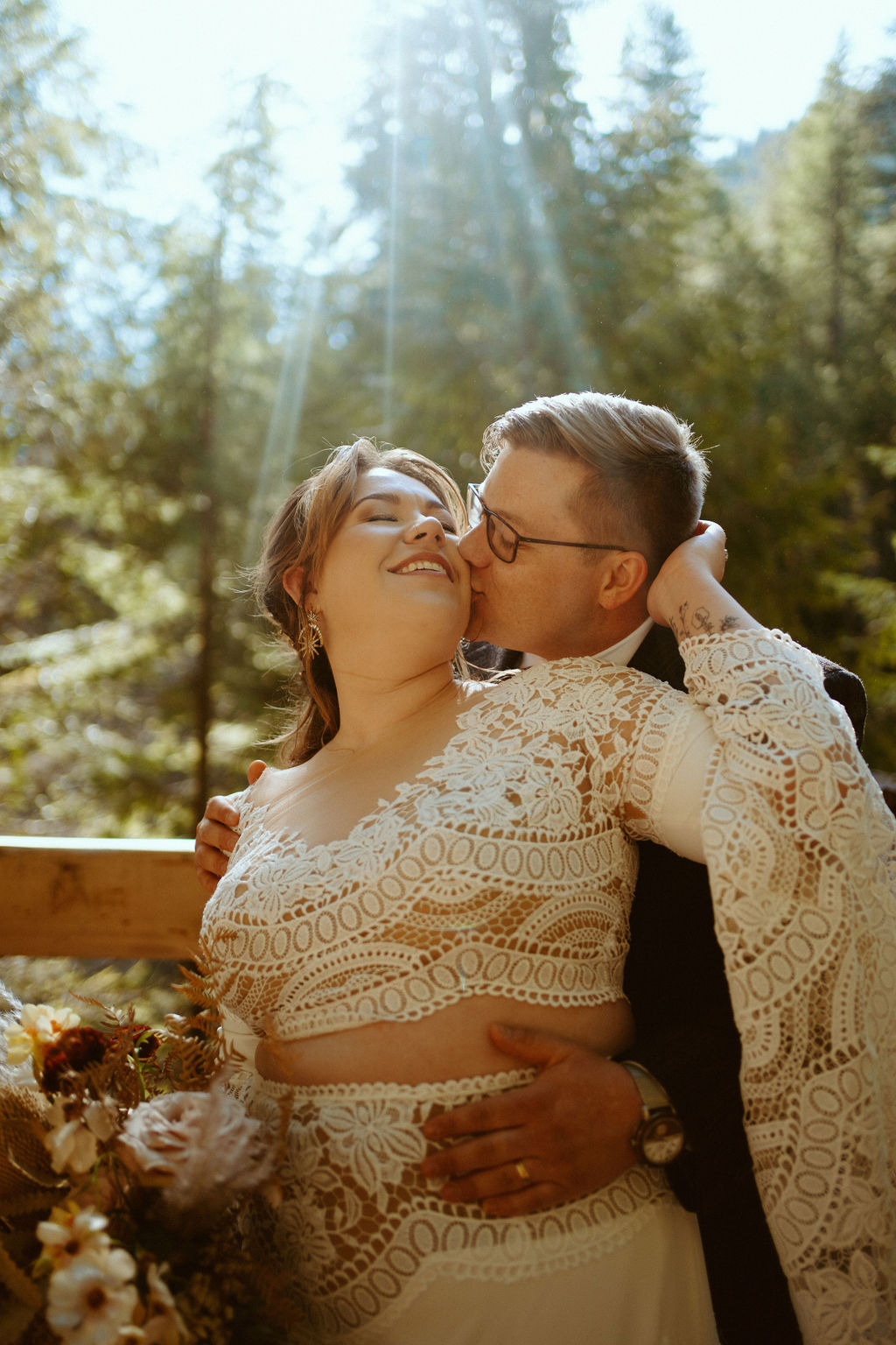 The groom is kissing the bride as she smiles and holds his head in the forest in front of a wooden fence 