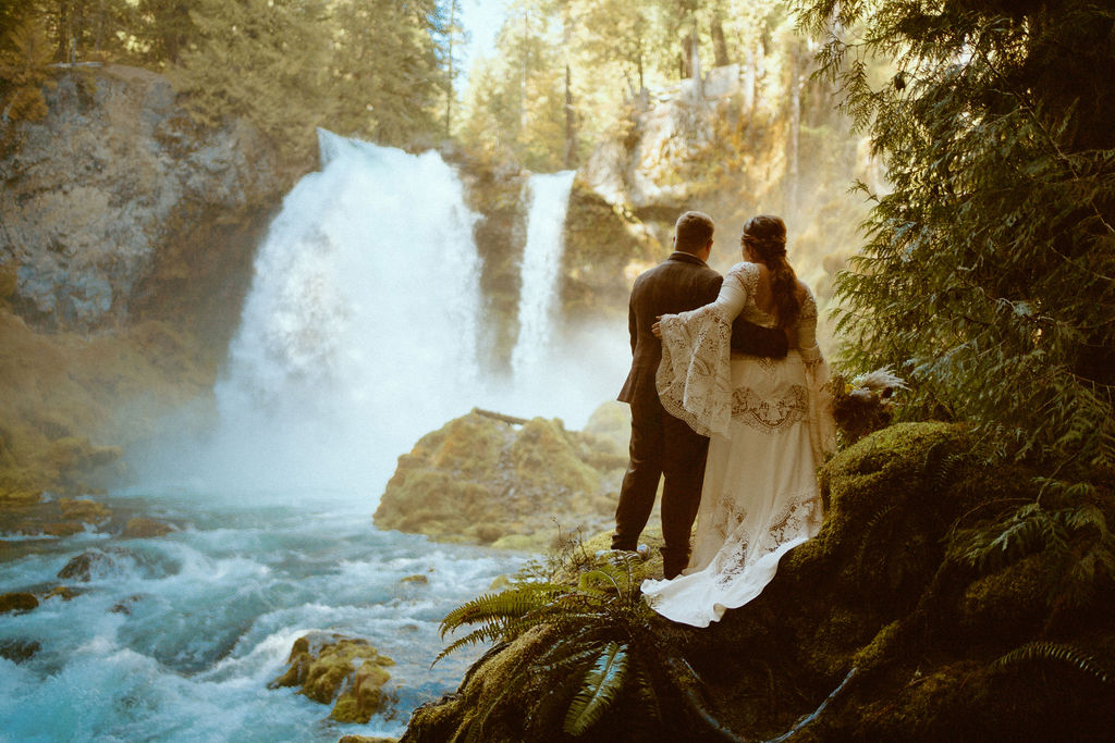Loloma Lodge Retro Whimsical Micro-Wedding. The bride in a long two piece white bohemian lace dress with the groom in a plaid suit looking out to the waterfall holding eachother