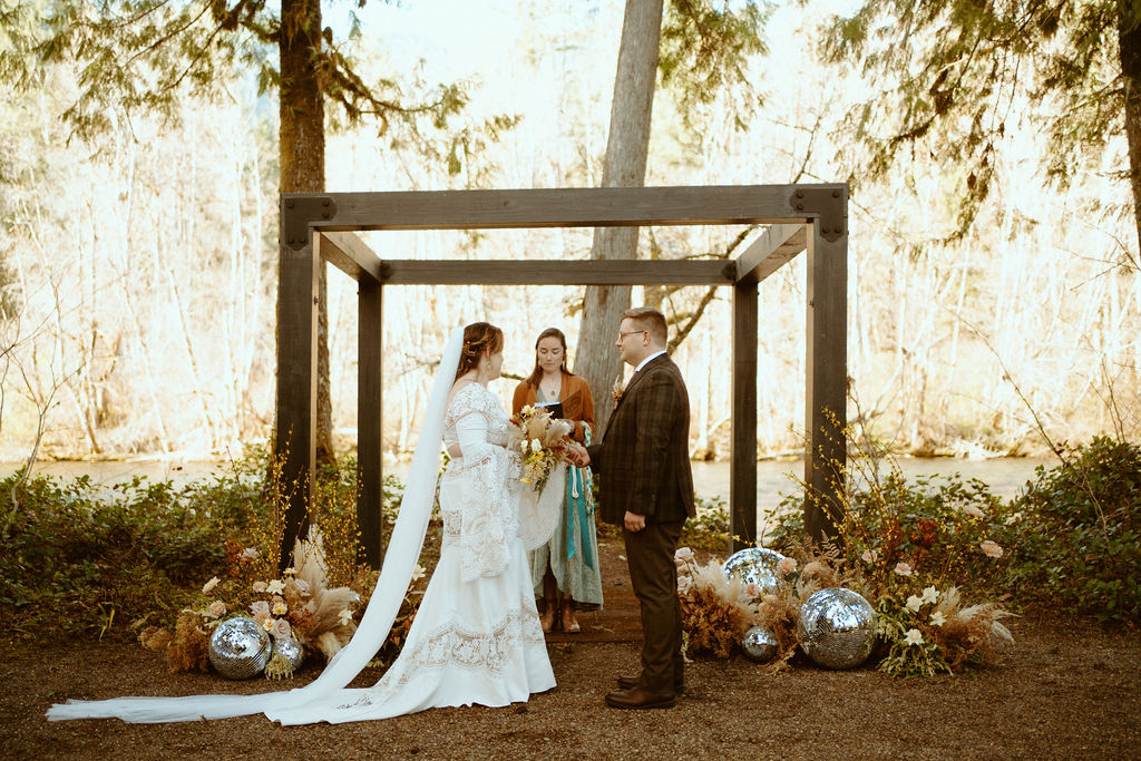 Loloma Lodge Retro Whimsical Micro-Wedding. Couple is standing in front of a wooden arch surrounded with wild flowers and disco balls while the officiant is reading.