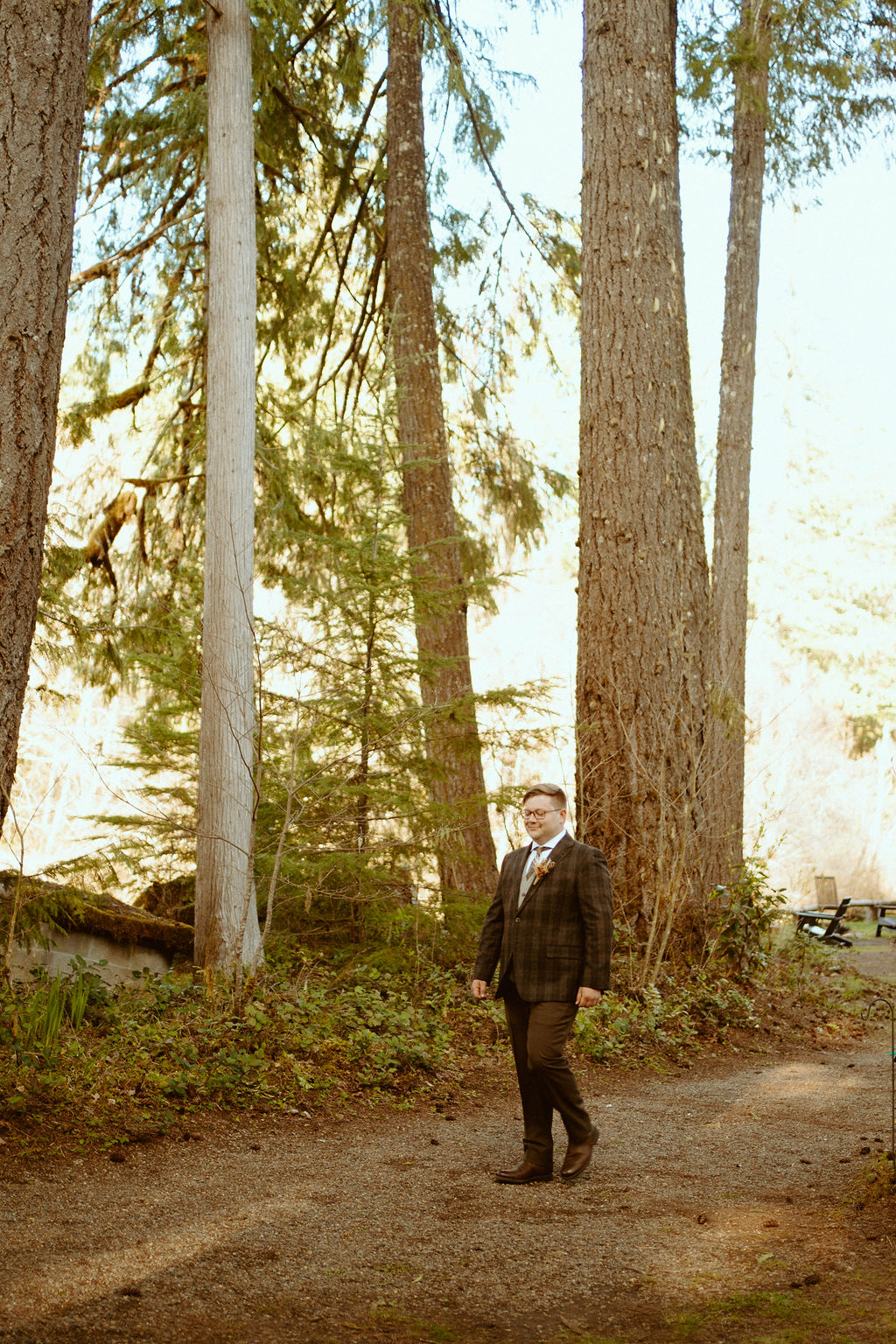 The groom walking down the aisle in between trees and greenery shrubs in a plaid suit with a small wildflower boutonniere 
