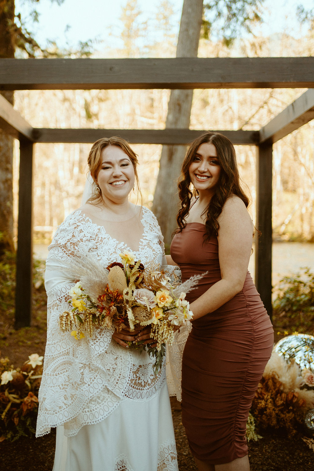 The bride poses with her maid of honor in front of the wooden arch 