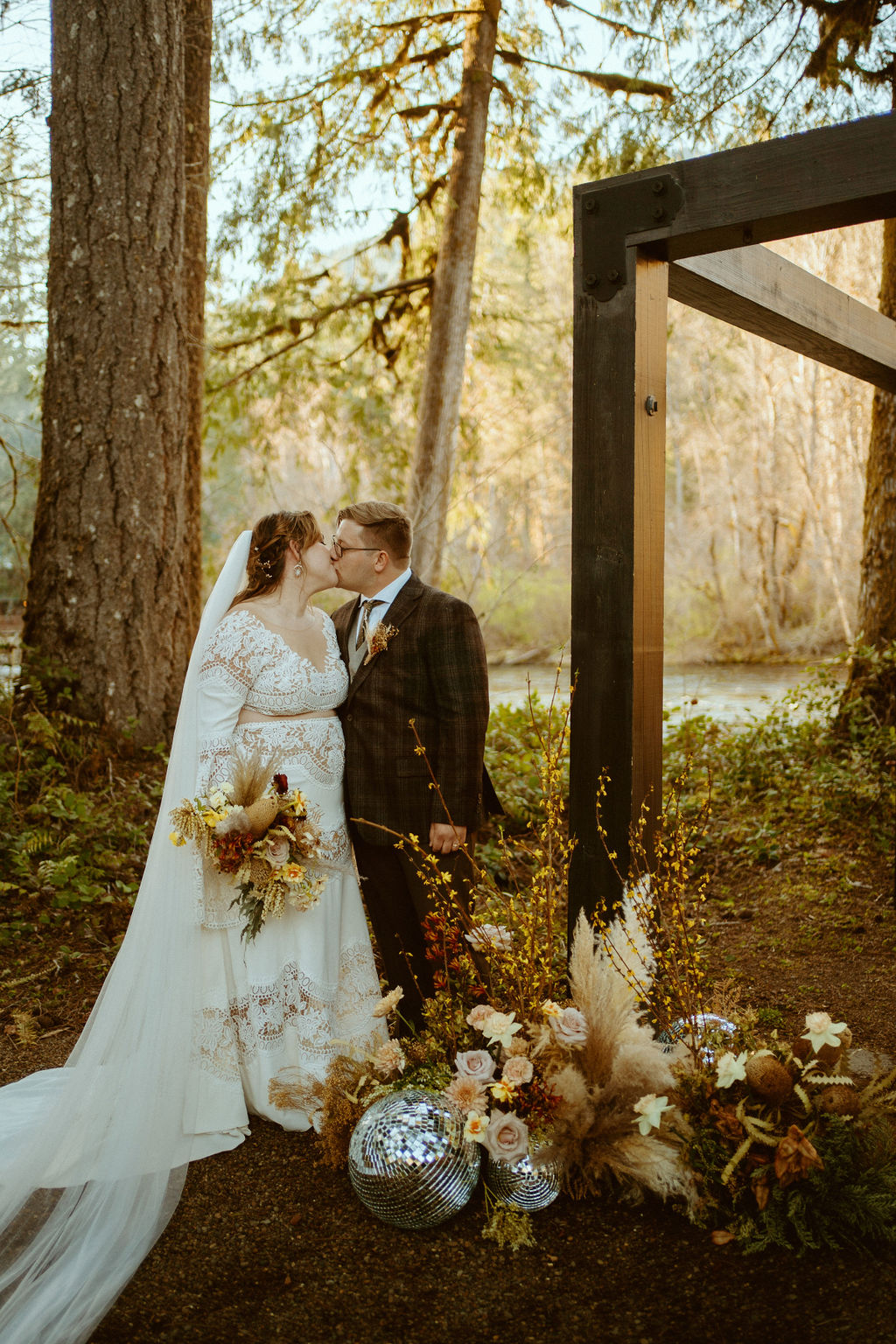 The bride and groom kiss to the side of the wooden arch standing behind the floral pieces and disco balls 
