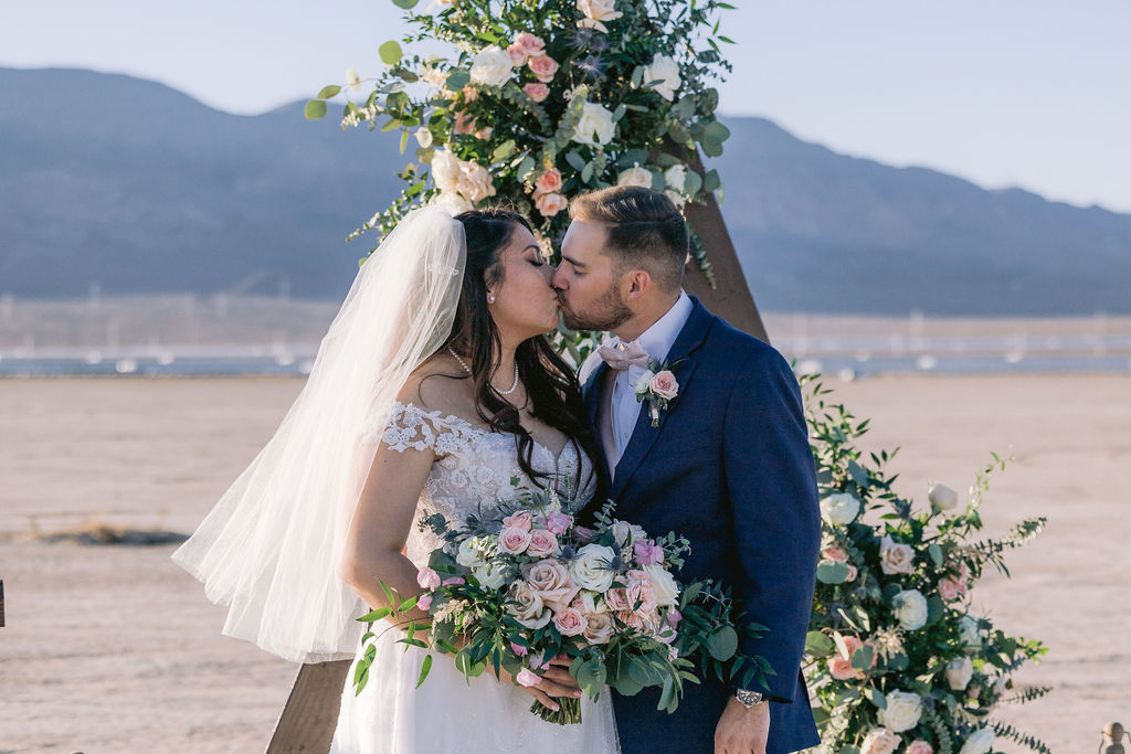 Navy Blue & Blush Pink Micro-Wedding Couple stands in front of their triangle wooden wedding arch sharing a kiss as she holds her blush pink floral bouquet. 