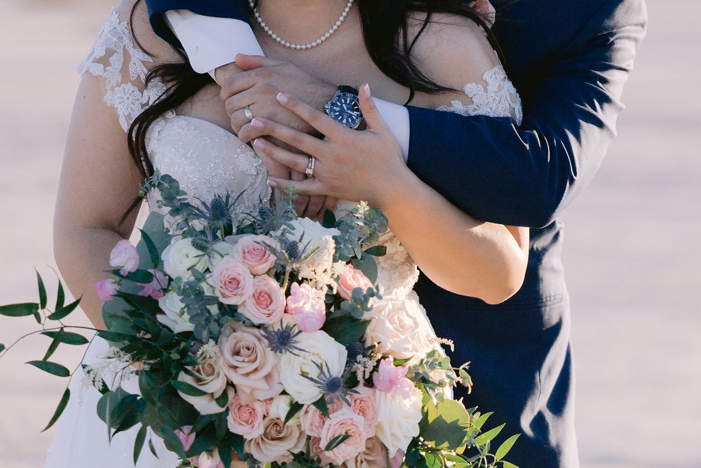 Close up of the grooms arms wrapped around the brides shoulders and her hand covers his as she holder her bouquet. 