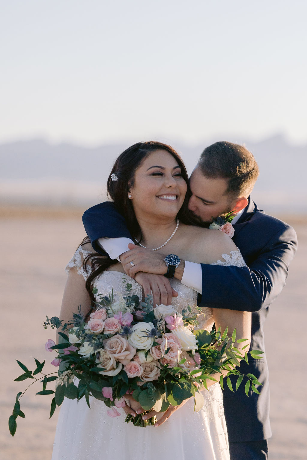 Navy Blue & Blush Pink Micro-Wedding groom wraps his arm around the brides shoulders and kisses her neck as she smiles holding her blush pink bouquet. 