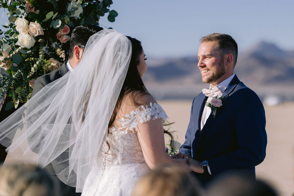 Navy Blue & Blush Pink Micro-Wedding groom smiles as he looks at his bride with her veil softly blowing in the wind. 