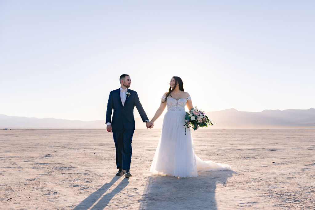 Navy Blue & Blush Pink Micro-Wedding walking hand in hand in the desert sunset. The sun adding the perfect glow behind them. 