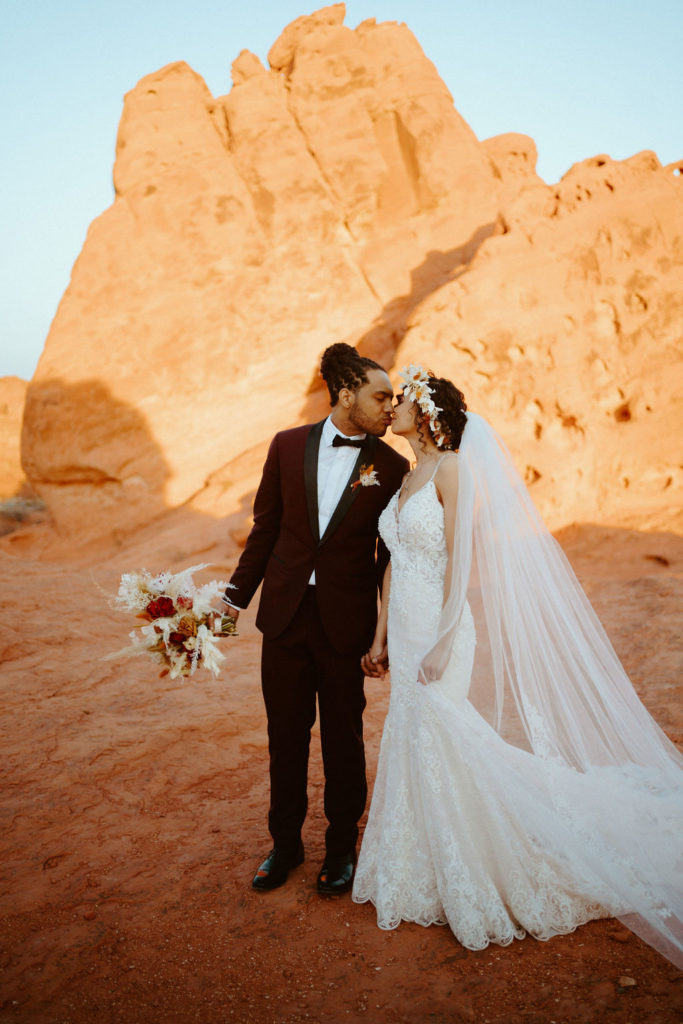 Bride and groom share a kiss as he holds her desert hues bouquet. 