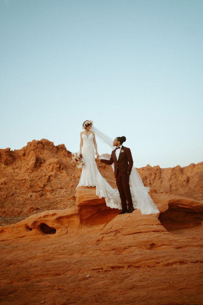 Romantic Mauve & Desert Hues Elopement bride standing and a rock formation the groom just bellow her holding hands. 
